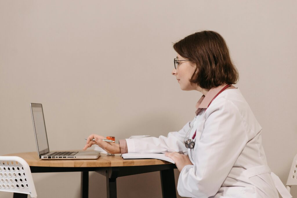 pexels-photo-5998487-5998487 Side profile of a female doctor in a white coat working on a laptop at a desk.